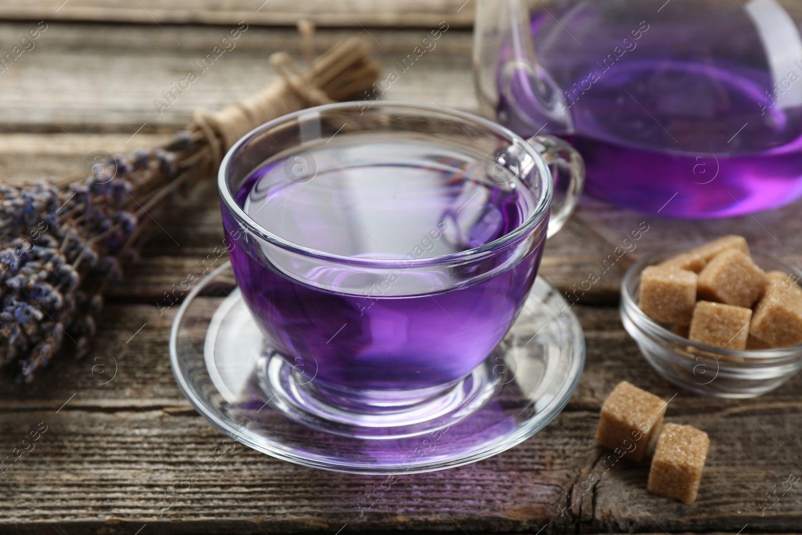 Photo of Aromatic lavender tea in glass cup, brown sugar and bunch of dry flowers on wooden table, closeup