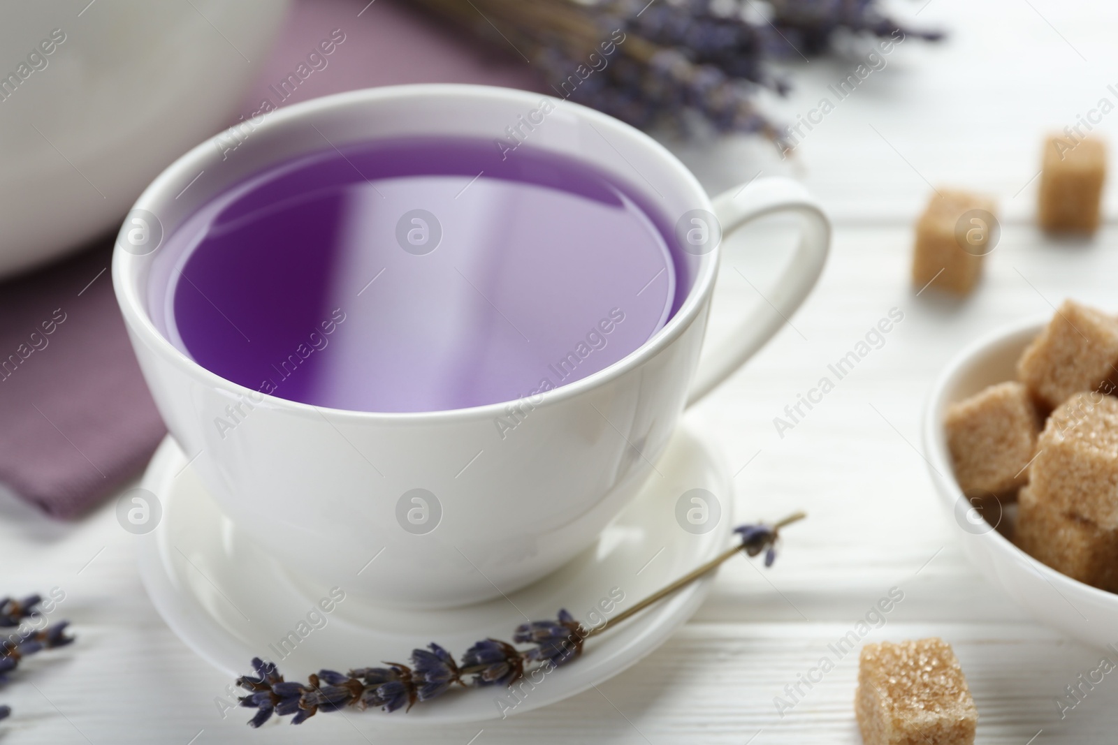 Photo of Aromatic lavender tea in cup, brown sugar and dry flowers on white wooden table, closeup