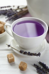 Photo of Aromatic lavender tea in cup, brown sugar and dry flowers on white wooden table, closeup