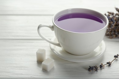 Photo of Aromatic lavender tea in cup, sugar cubes and dry flowers on white wooden table, closeup. Space for text