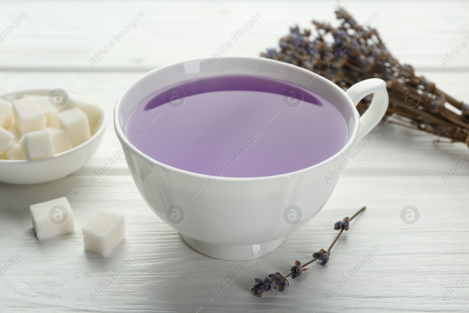 Photo of Aromatic lavender tea in cup, sugar cubes and dry flowers on white wooden table, closeup