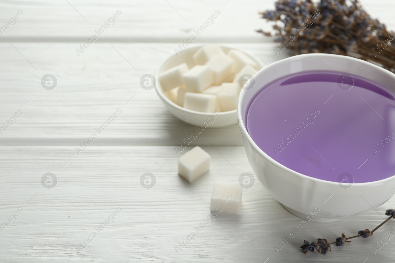 Photo of Aromatic lavender tea in cup, sugar cubes and dry flowers on white wooden table, closeup. Space for text