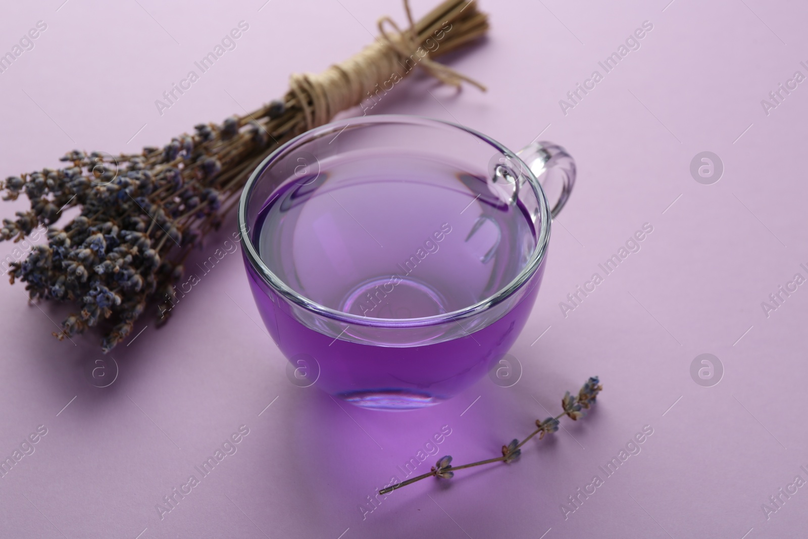 Photo of Aromatic lavender tea in glass cup and bunch of dry flowers on lilac background, closeup