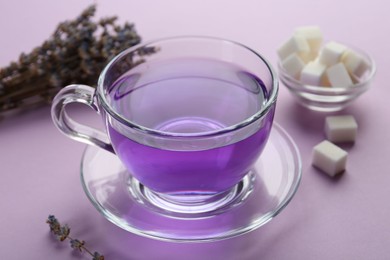 Photo of Aromatic lavender tea in glass cup, sugar cubes and dry flowers on lilac background, closeup