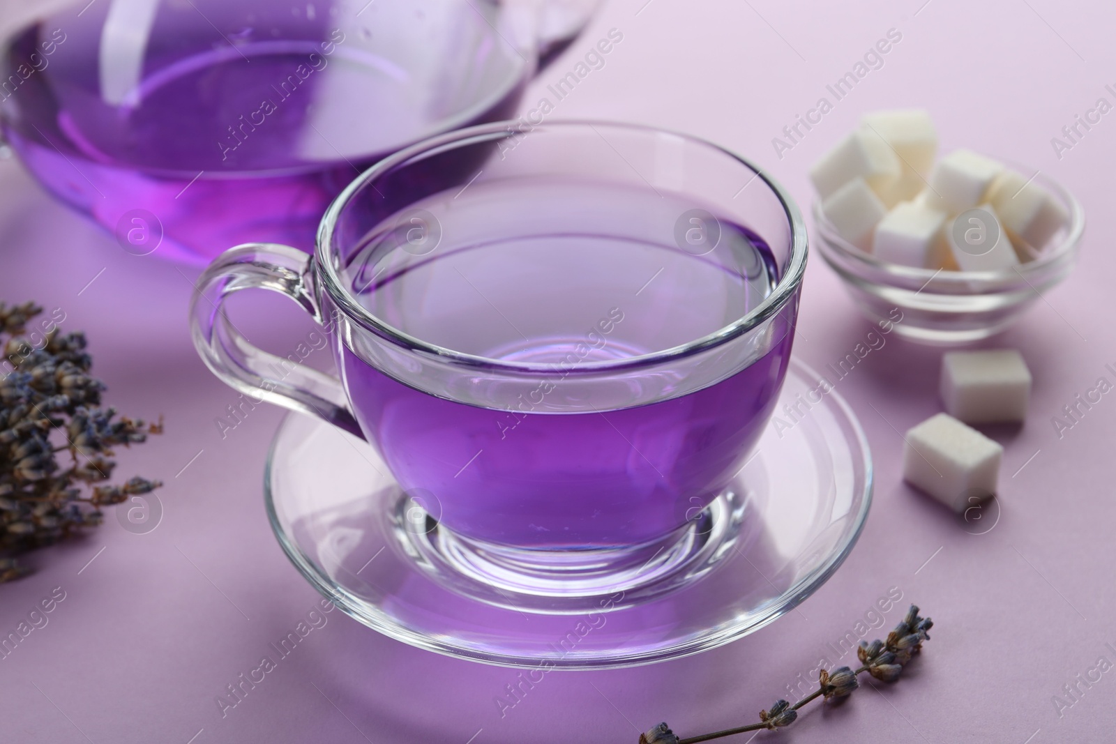 Photo of Aromatic lavender tea in glass cup, sugar cubes and dry flowers on lilac background, closeup