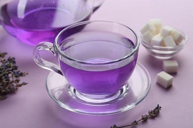 Aromatic lavender tea in glass cup, sugar cubes and dry flowers on lilac background, closeup