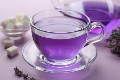 Photo of Aromatic lavender tea in glass cup, sugar cubes and dry flowers on lilac background, closeup