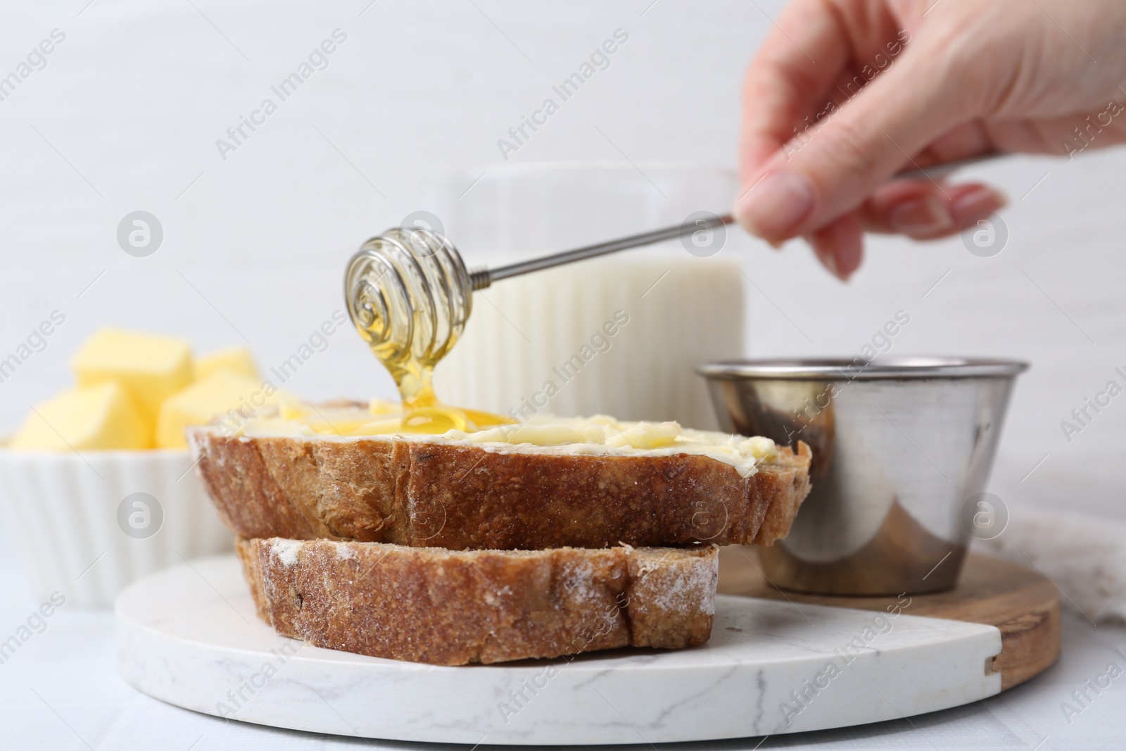 Photo of Woman pouring honey onto slices of bread with butter at white table, closeup