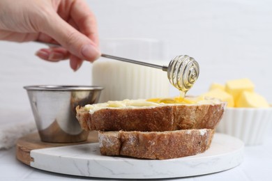 Woman pouring honey onto slices of bread with butter at white table, closeup