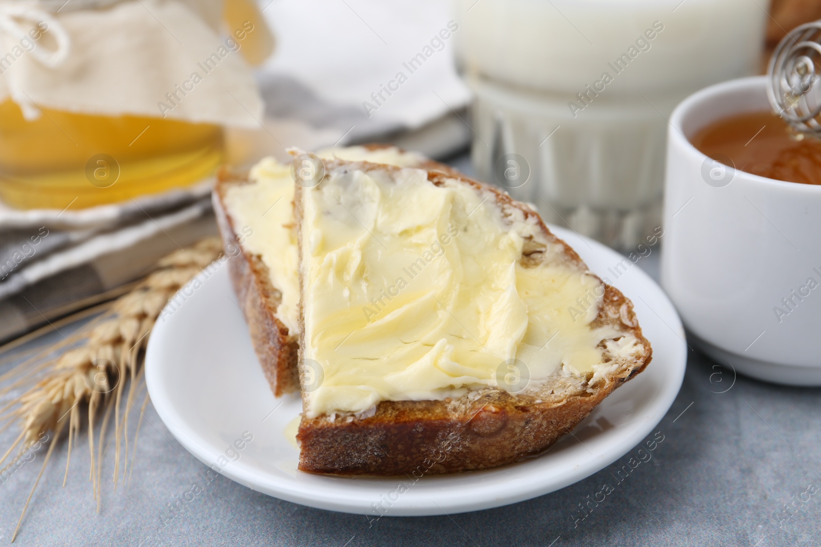 Photo of Slices of bread with butter, honey, milk and spike on light grey table, closeup