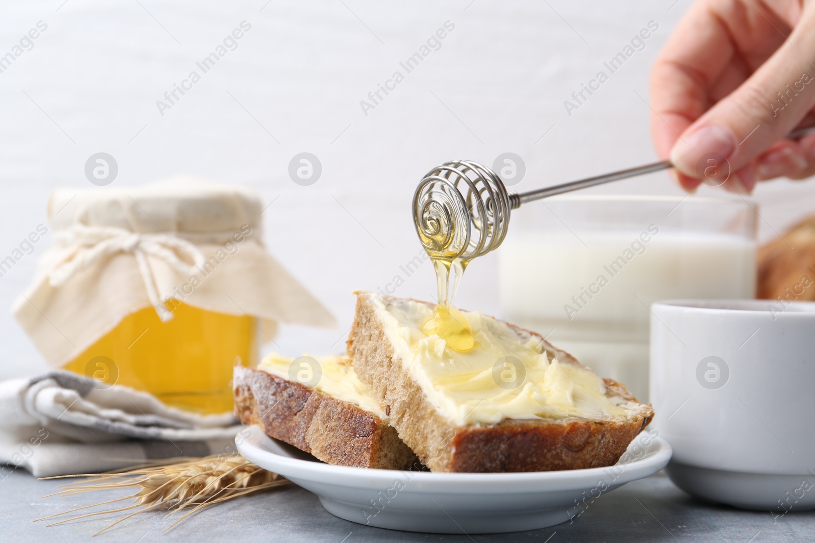 Photo of Woman pouring honey onto slices of bread with butter at grey table, closeup