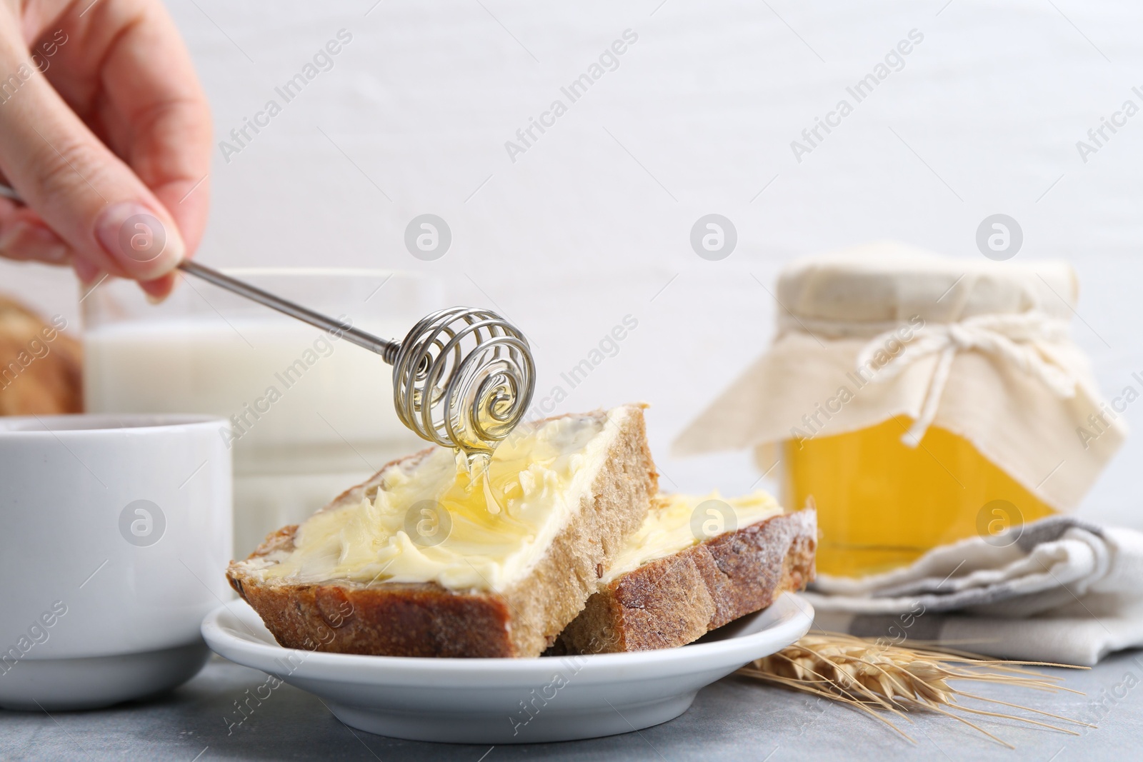 Photo of Woman pouring honey onto slices of bread with butter at grey table, closeup