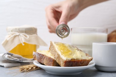 Photo of Woman pouring honey onto slices of bread with butter at grey table, closeup