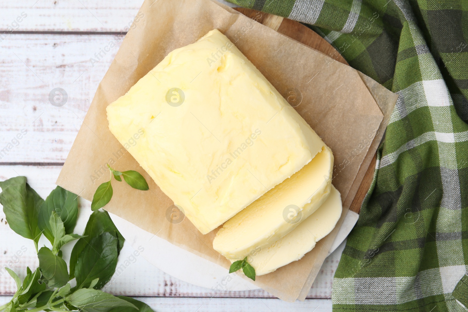 Photo of Cut brick of butter and basil on white wooden table, flat lay