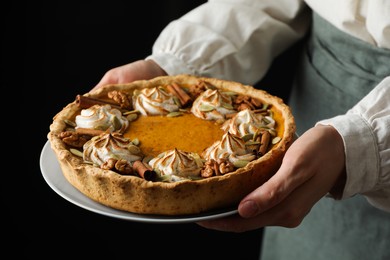 Photo of Woman holding homemade pumpkin pie with whipped cream, seeds and cinnamon on black background, closeup