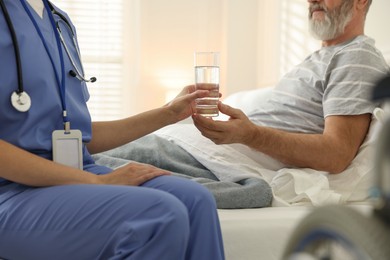 Photo of Caregiver giving water to senior man on bed indoors, closeup. Home health care service