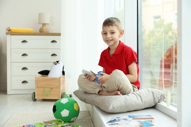 Creating vision board. Little boy cutting out picture near window indoors