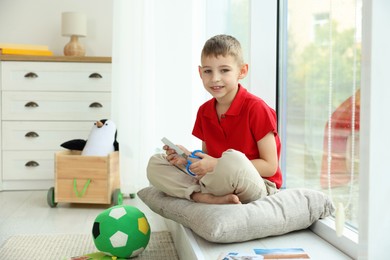 Creating vision board. Little boy cutting out picture near window indoors