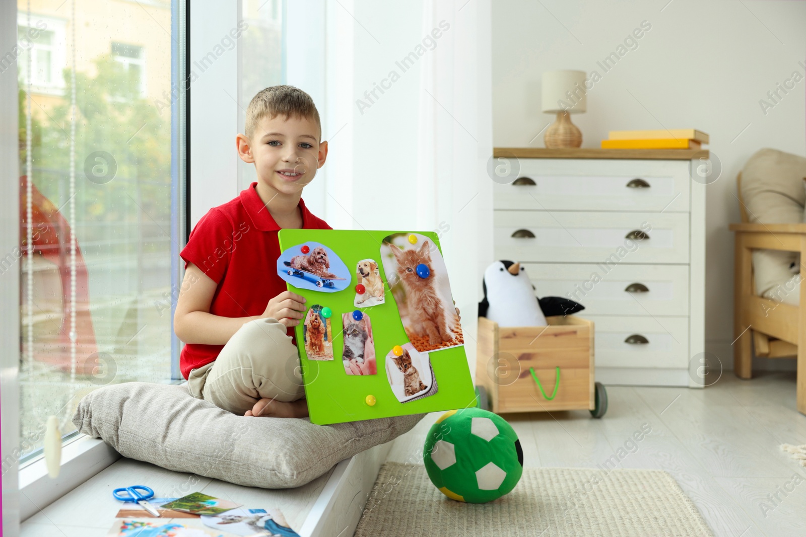 Photo of Little boy holding vision board with different pictures near window indoors