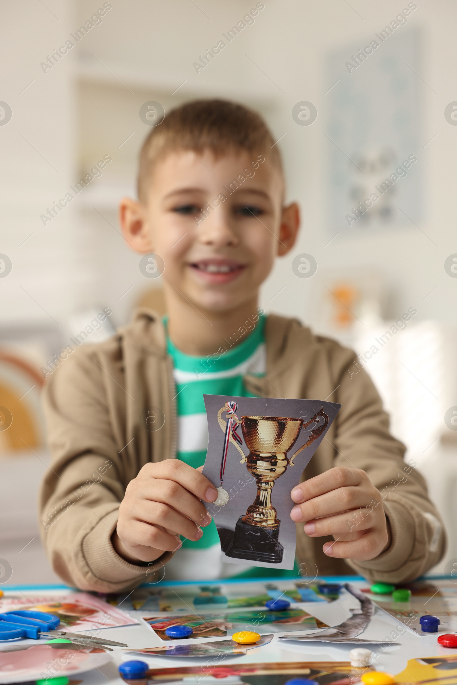 Photo of Creating vision board. Little boy with picture of golden cup trophy at table indoors