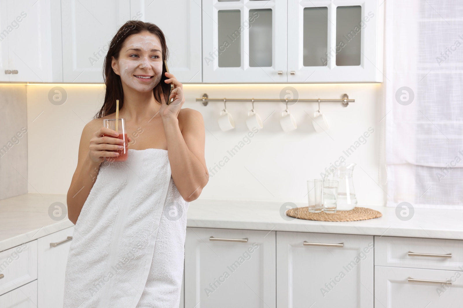 Photo of Spa day. Beautiful woman with face mask and juice talking on smartphone in kitchen, space for text