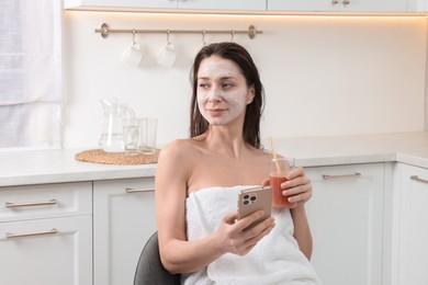 Spa day. Beautiful woman with face mask and juice using smartphone in kitchen