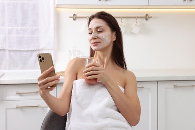 Spa day. Beautiful woman with face mask and juice using smartphone in kitchen