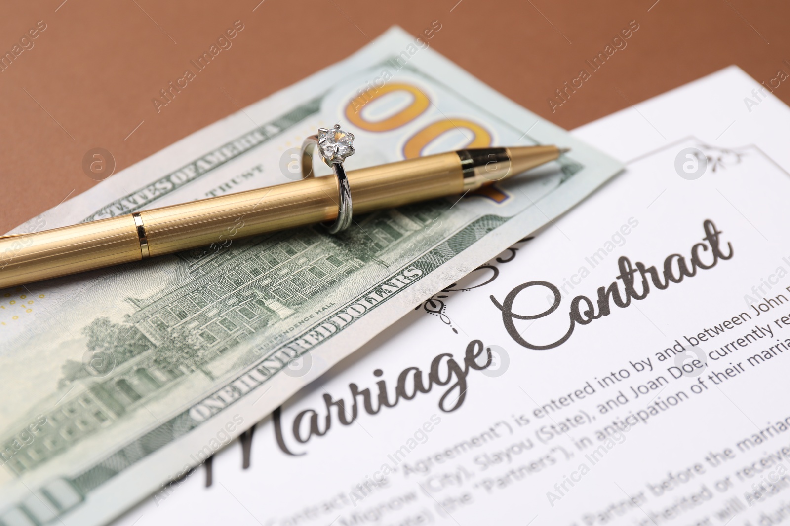 Photo of Marriage contract, pen, money and ring on brown background, closeup