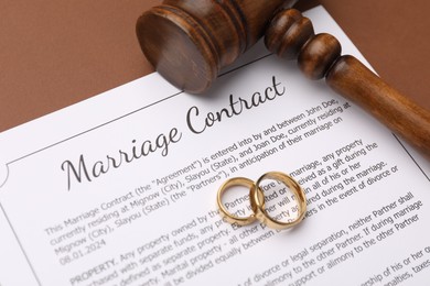 Photo of Marriage contract, gavel and golden rings on brown background, closeup