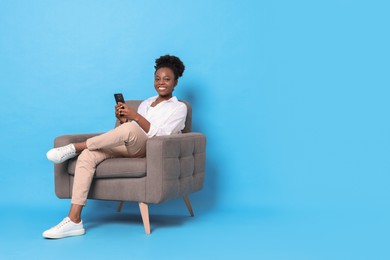 Smiling woman with smartphone sitting in armchair on light blue background. Space for text