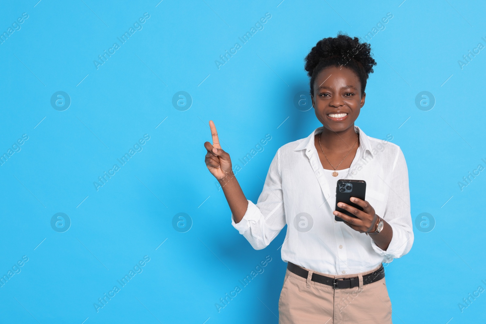 Photo of Cheerful woman with smartphone pointing at something on light blue background. Space for text