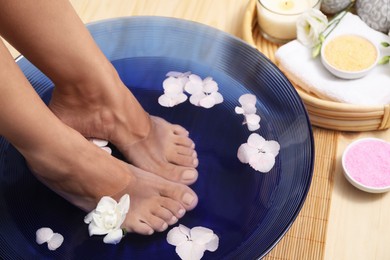 Photo of Woman soaking her feet in bowl with water and flowers on floor, closeup. Spa treatment