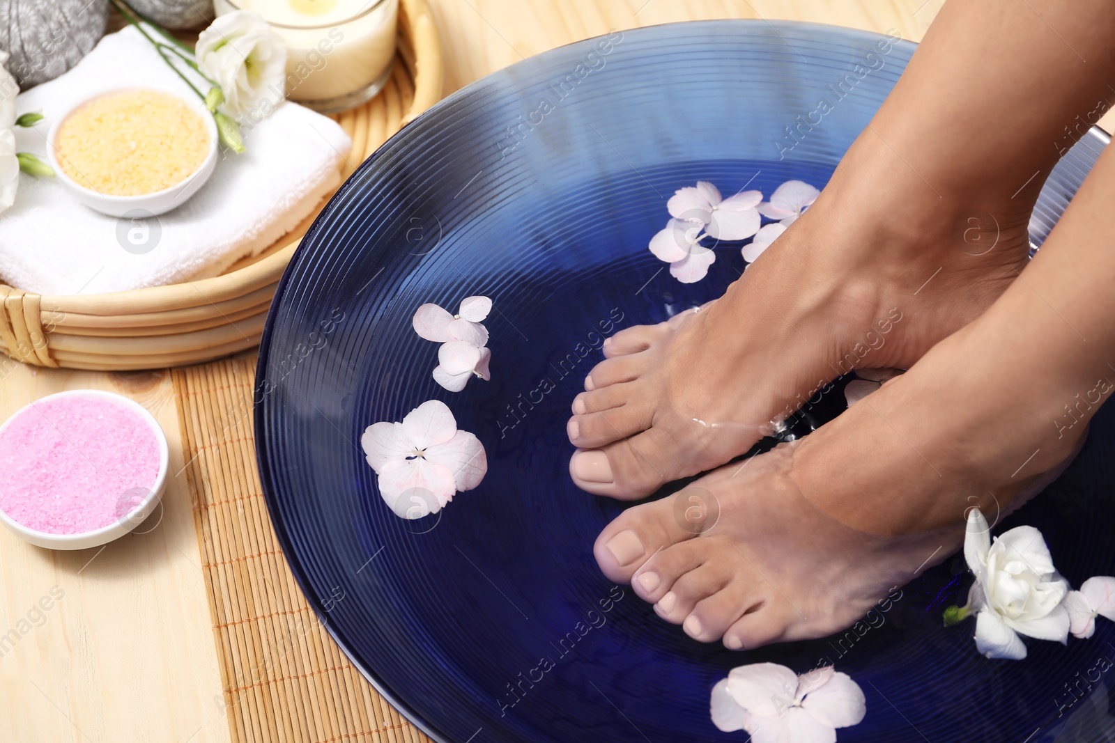 Photo of Woman soaking her feet in bowl with water and flowers on floor, closeup. Spa treatment