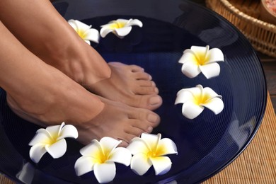 Photo of Woman soaking her feet in bowl with water and plumeria flowers on floor, closeup. Spa treatment