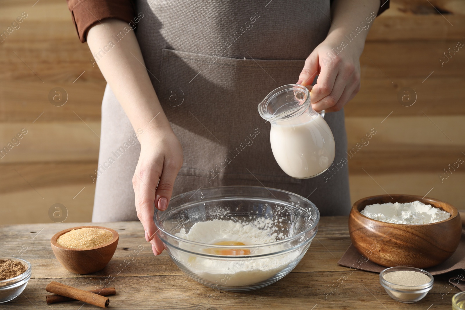 Photo of Making cinnamon rolls. Woman pouring milk into bowl at wooden table, closeup