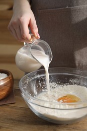 Photo of Making cinnamon rolls. Woman pouring milk into bowl at wooden table, closeup