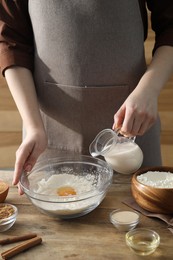 Photo of Making cinnamon rolls. Woman pouring milk into bowl at wooden table, closeup