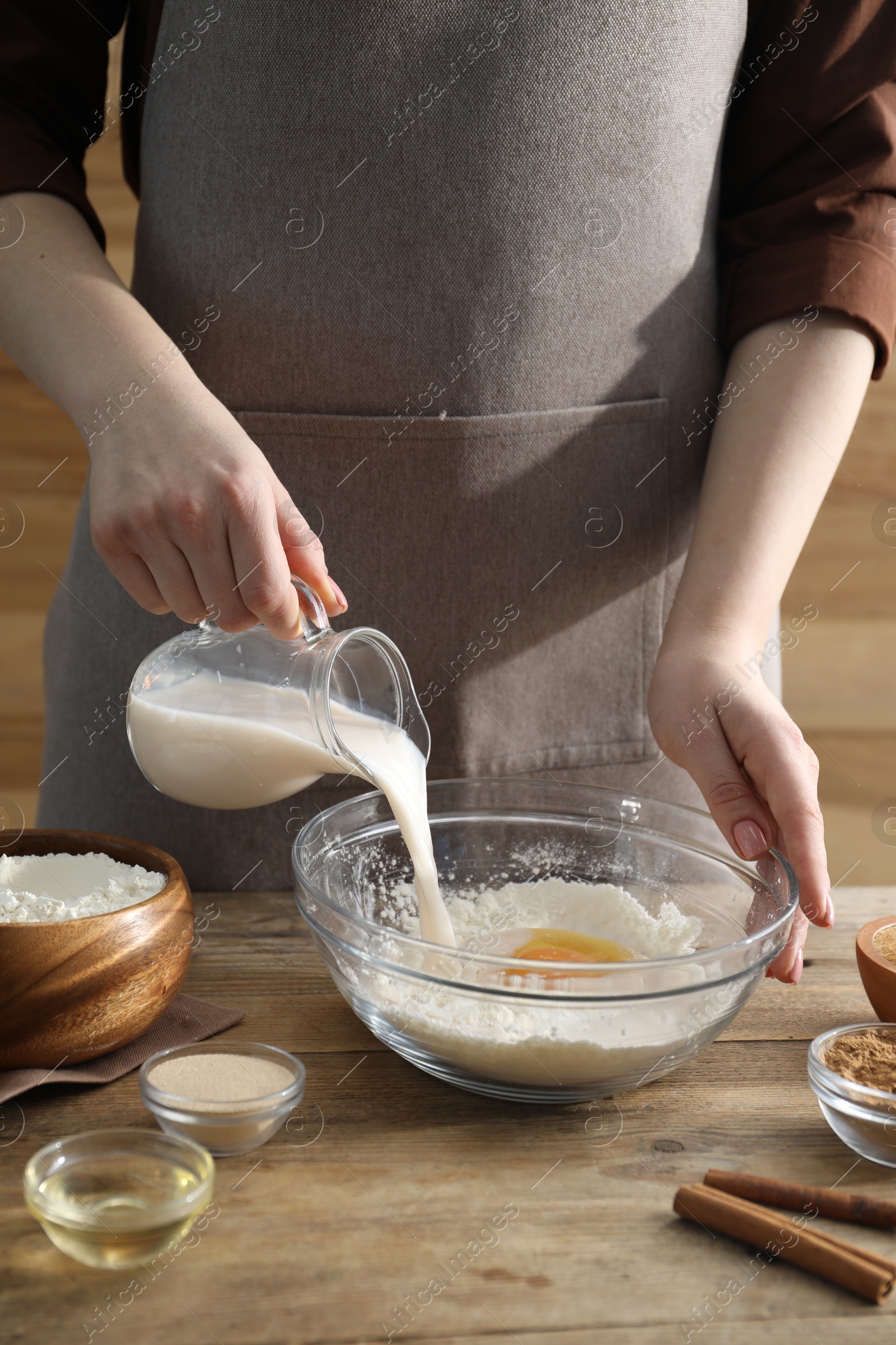 Photo of Making cinnamon rolls. Woman pouring milk into bowl at wooden table, closeup