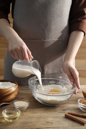 Photo of Making cinnamon rolls. Woman pouring milk into bowl at wooden table, closeup