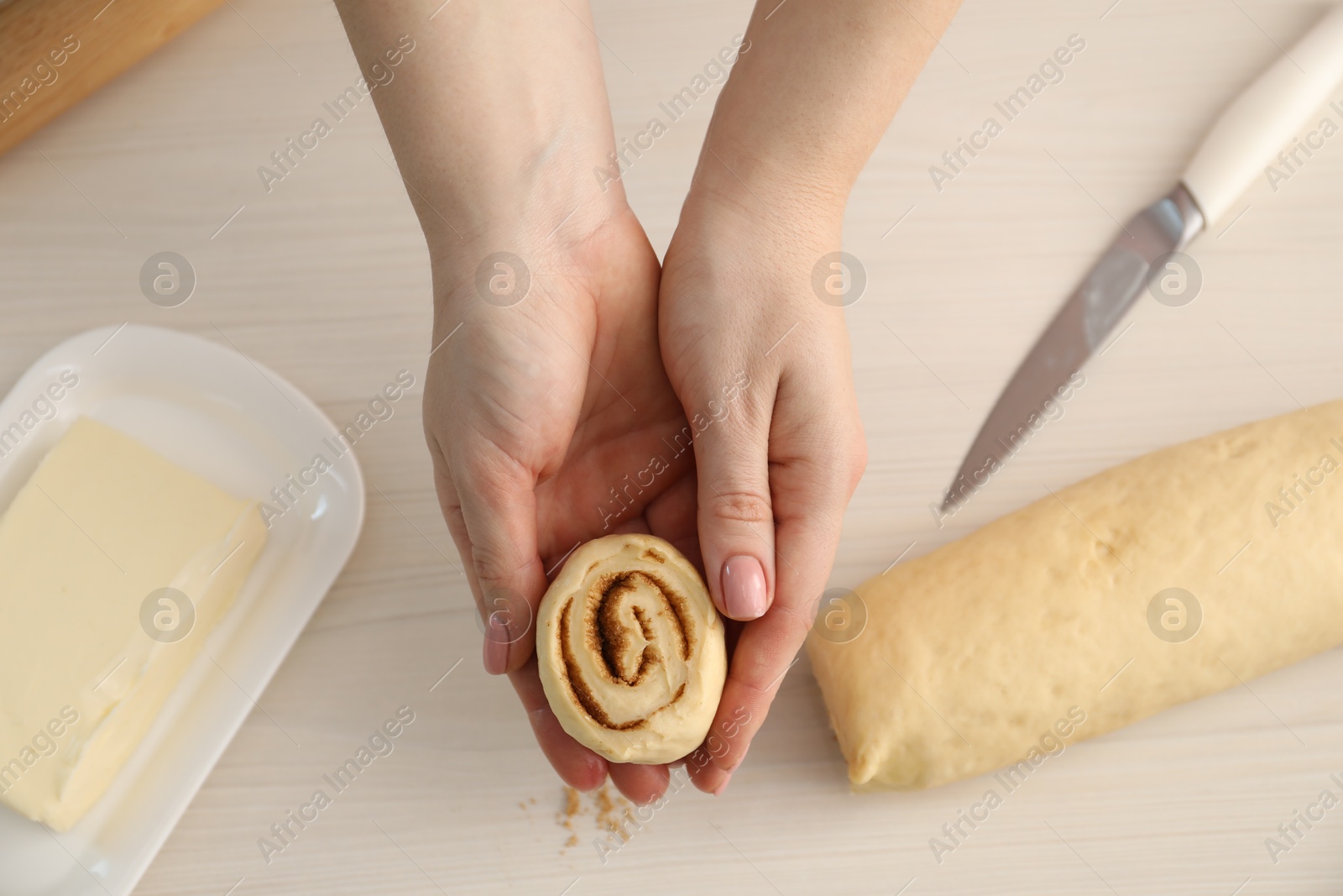 Photo of Woman with uncooked cinnamon roll at white wooden table, top view