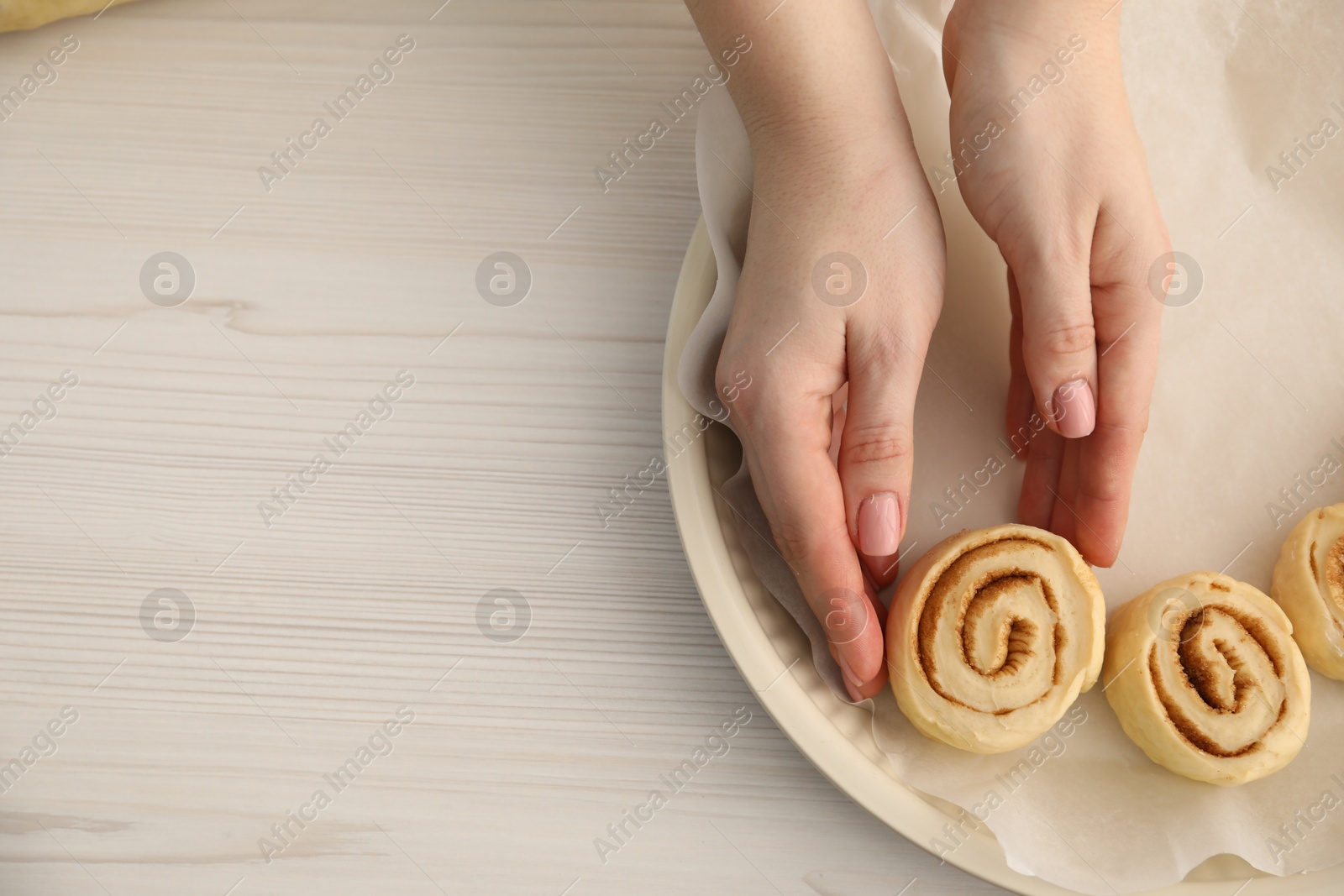 Photo of Woman putting cinnamon roll into baking dish at white wooden table, closeup. Space for text