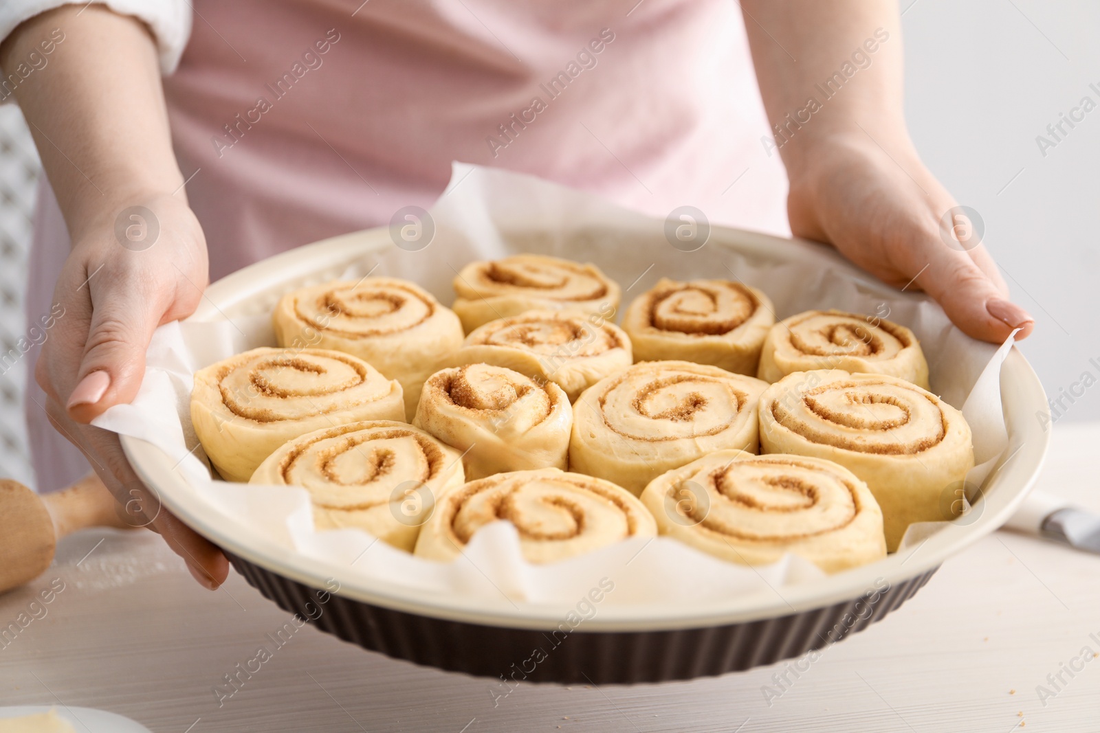 Photo of Woman holding baking dish with uncooked cinnamon rolls at white table, closeup