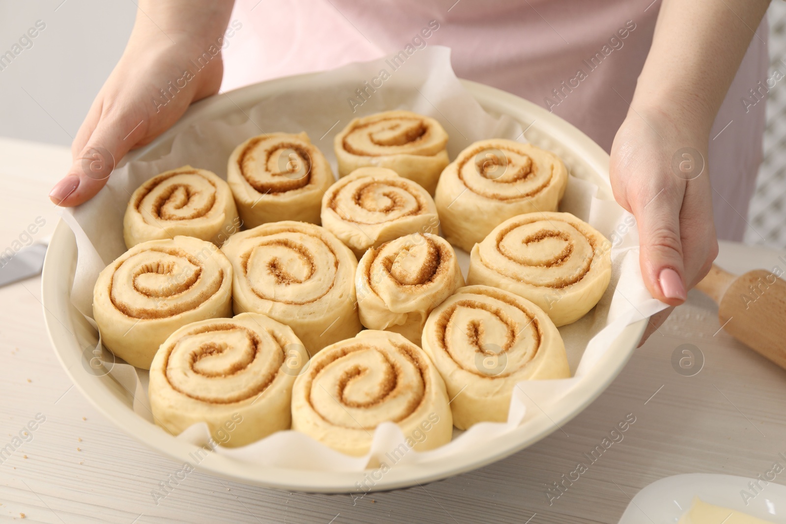 Photo of Woman holding baking dish with uncooked cinnamon rolls at wooden table, closeup