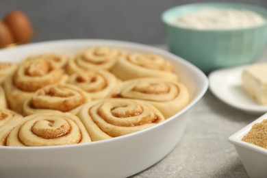 Photo of Raw cinnamon rolls in baking dish and ingredients on grey table, closeup