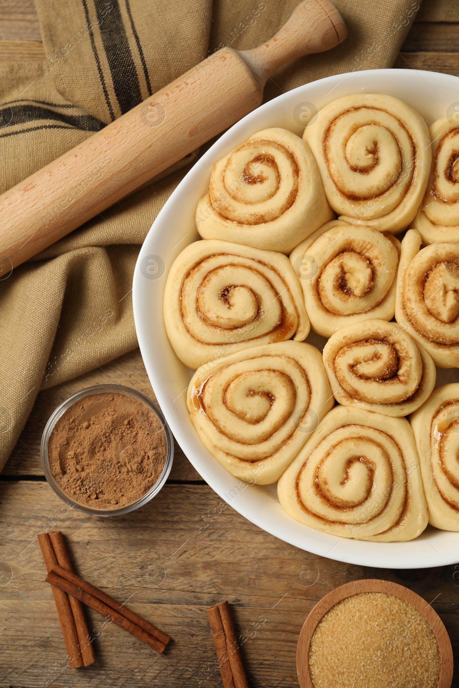 Photo of Raw cinnamon rolls in baking dish, rolling pin, cocoa powder and brown sugar on wooden table, flat lay