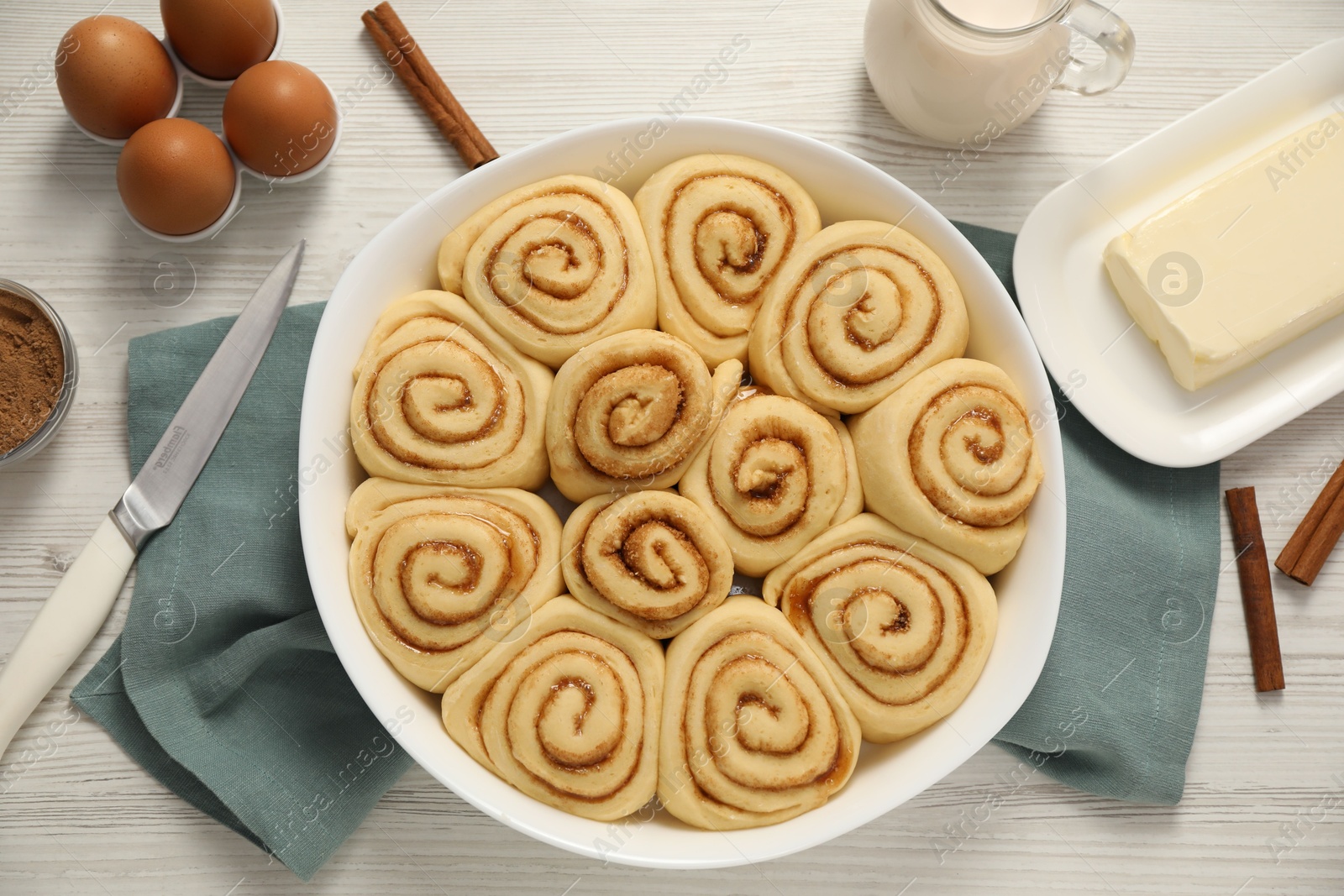 Photo of Raw cinnamon rolls and different ingredients on light wooden table, flat lay