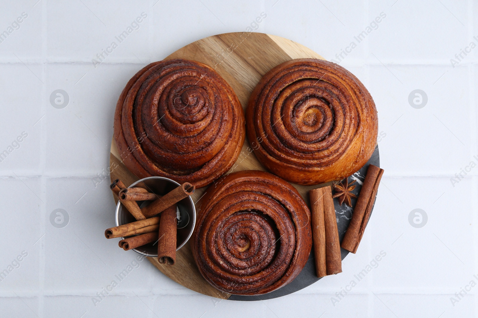 Photo of Delicious cinnamon roll buns and spices on white tiled table, top view