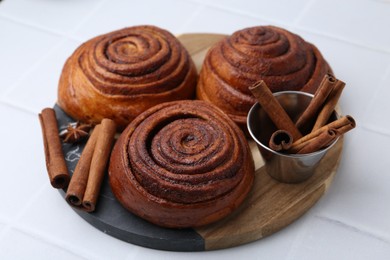 Photo of Delicious cinnamon roll buns and spices on white tiled table, closeup