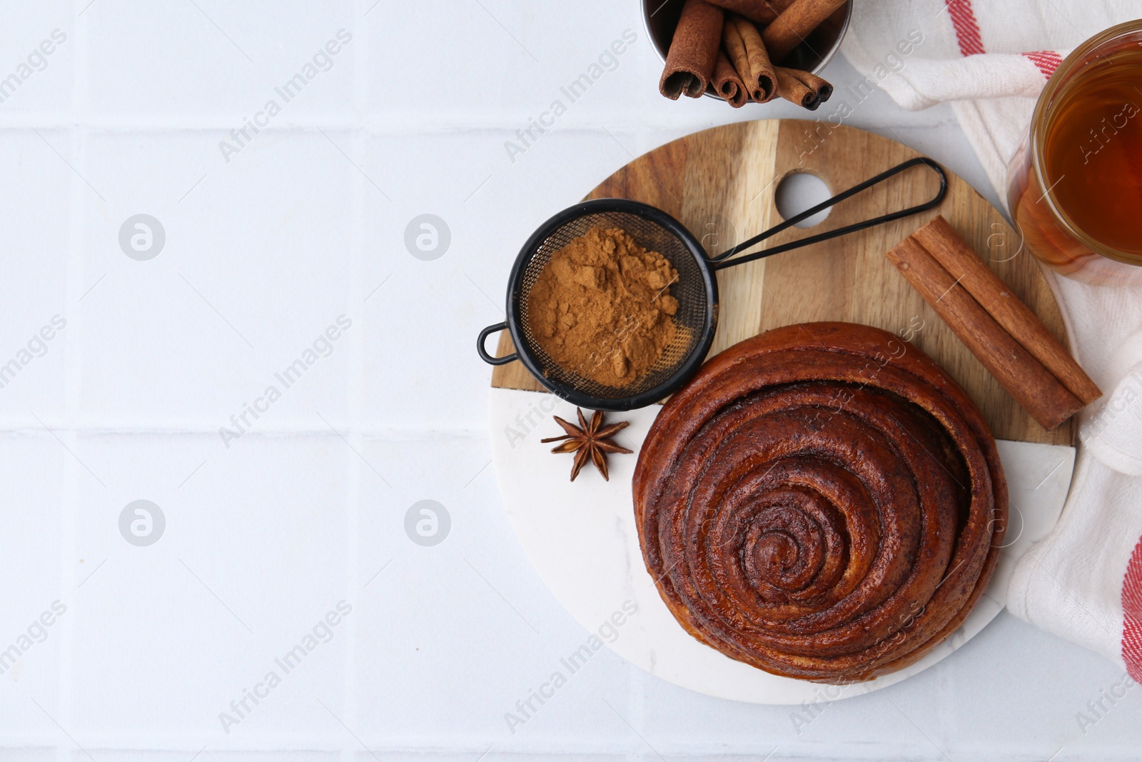 Photo of Delicious cinnamon roll bun, tea and spices on white tiled table, flat lay. Space for text