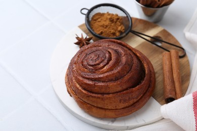 Photo of Delicious cinnamon roll bun and spices on white tiled table, closeup